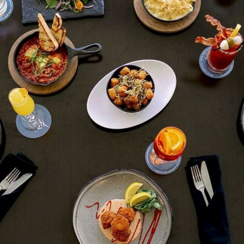 The image shows a table set with various brunch dishes and drinks, including waffles, cocktails, fried food, and appetizers, along with utensils.