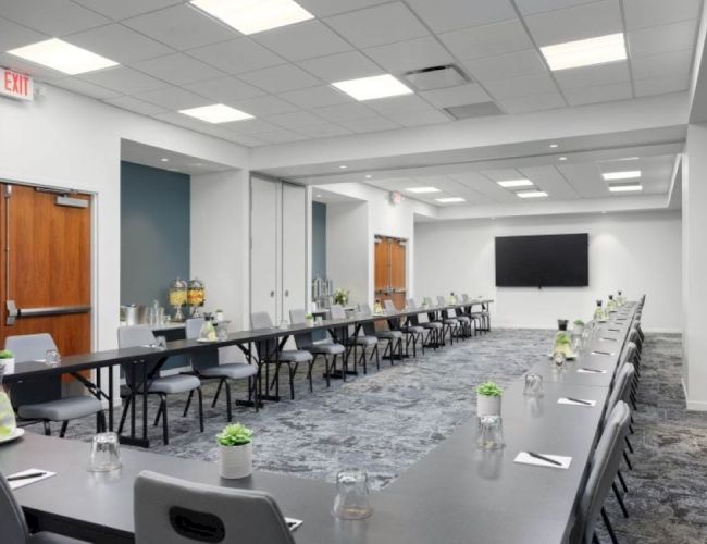 A modern conference room with long tables arranged in a U-shape, equipped with chairs, water bottles, and potted plants. Large screen on the wall.