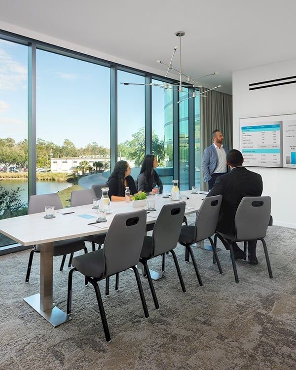 A conference room with four people, a large table, chairs, a presentation screen displaying charts, and large windows showcasing an outdoor view.