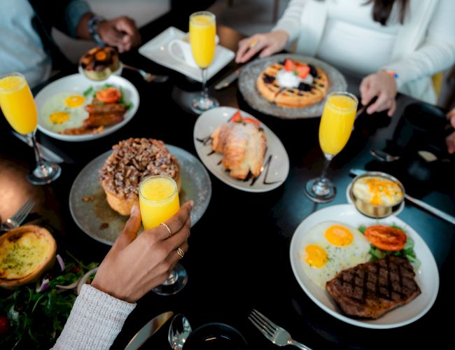 A table with brunch dishes and glasses of orange juice, including steak, eggs, waffles, pastries, and a salad, surrounded by people reaching in.