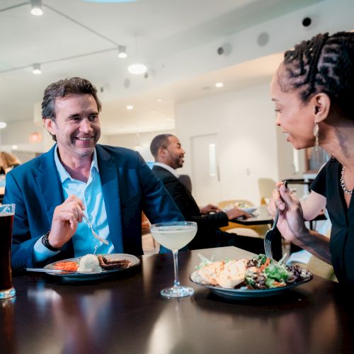 Two people are dining at a restaurant, enjoying a meal and drinks, with other guests visible in the background.