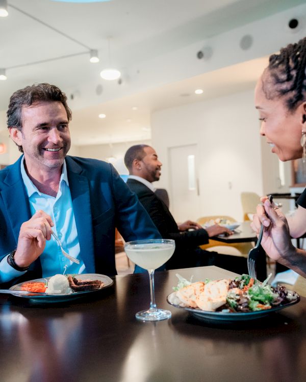 Two people are dining at a restaurant, enjoying a meal and drinks, with other guests visible in the background.