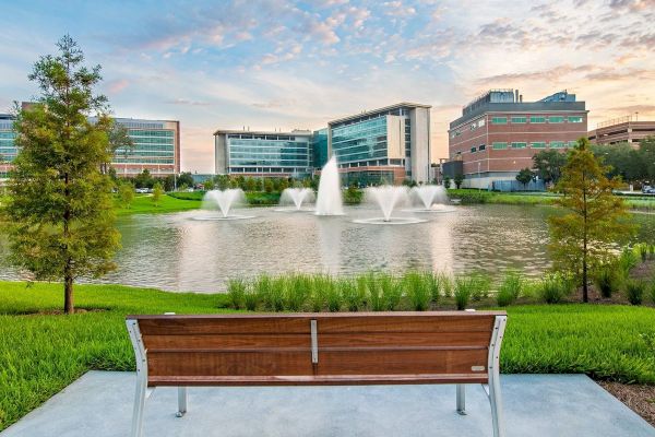 A wooden bench facing a pond with fountains, surrounded by greenery, with modern buildings in the background under a partly cloudy sky.