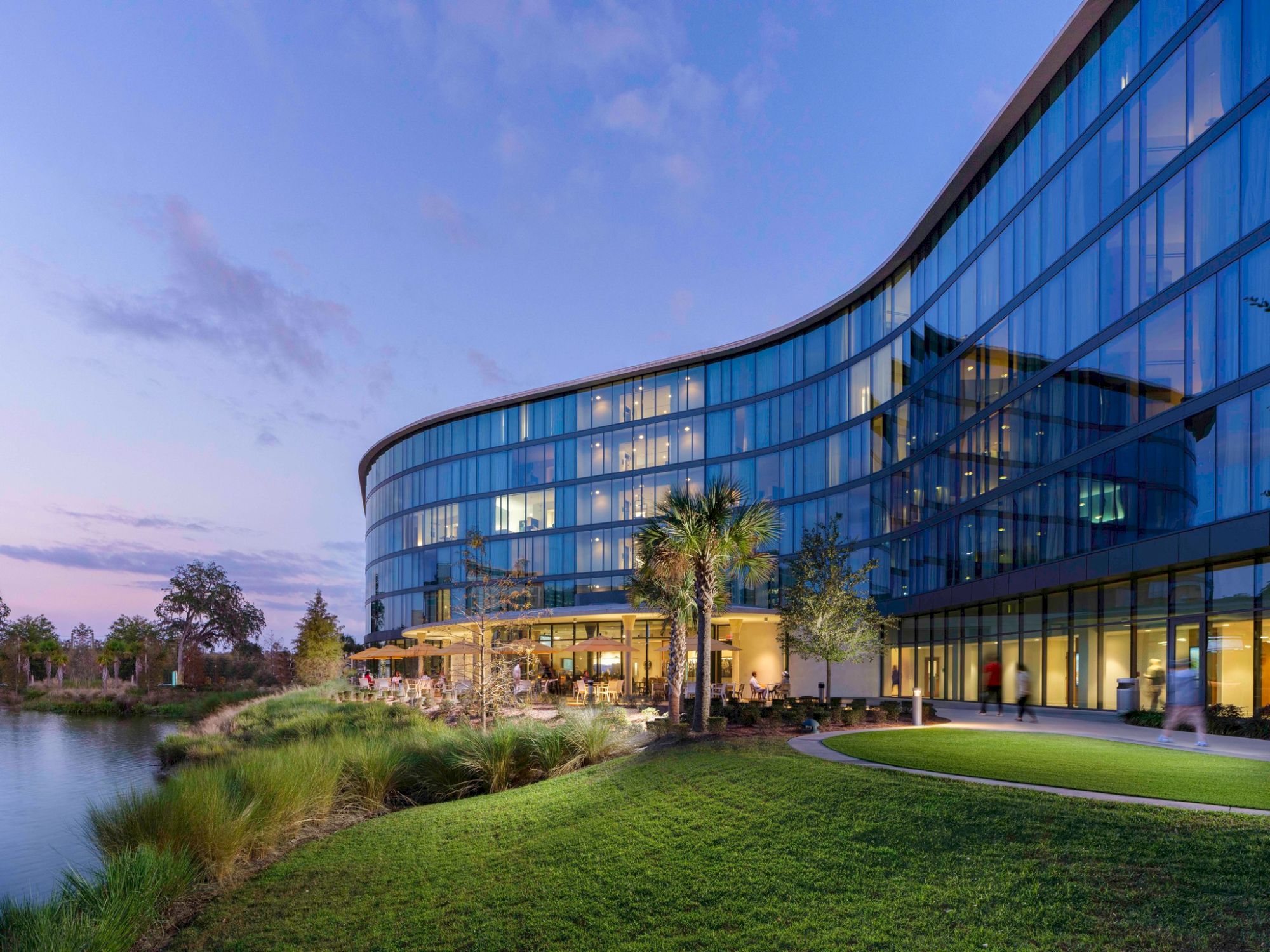 The image shows a modern, multi-story glass building with a curved design, situated beside a pond and surrounded by green landscaping and trees.