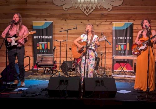 Three musicians perform on a stage with a wooden backdrop, flanked by banners that read 