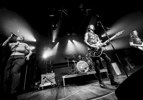 A black and white photo of a band performing live on stage, with musicians playing instruments under dramatic lighting.