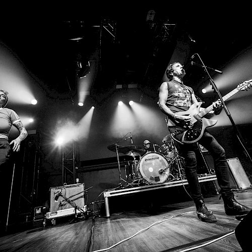 A black and white photo of a band performing live on stage, with musicians playing instruments under dramatic lighting.