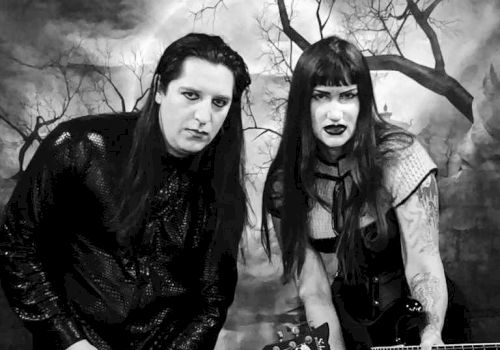 Two people in gothic attire, holding guitars, pose against a dark, eerie background with barren trees.