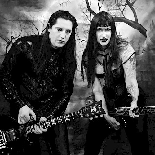 Two people in gothic attire, holding guitars, pose against a dark, eerie background with barren trees.