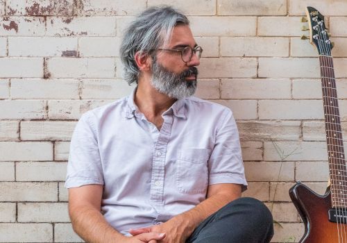 A man with gray hair and a beard sits against a brick wall, looking to his left. Beside him is an electric guitar resting against the wall.