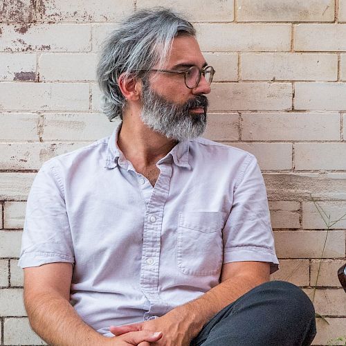 A man with gray hair and a beard sits against a brick wall, looking to his left. Beside him is an electric guitar resting against the wall.