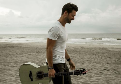 A person is walking barefoot on a beach, holding an acoustic guitar, with the ocean and a cloudy sky in the background.