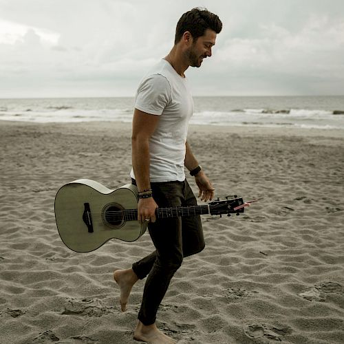 A person is walking barefoot on a beach, holding an acoustic guitar, with the ocean and a cloudy sky in the background.