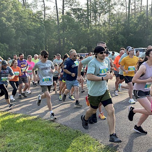 A group of people are running together in an outdoor setting, likely participating in a race or marathon, with trees and greenery in the background.