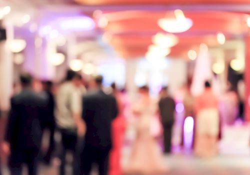 The image shows a blurred view of people attending a formal event or gathering in a well-lit, elegant venue.