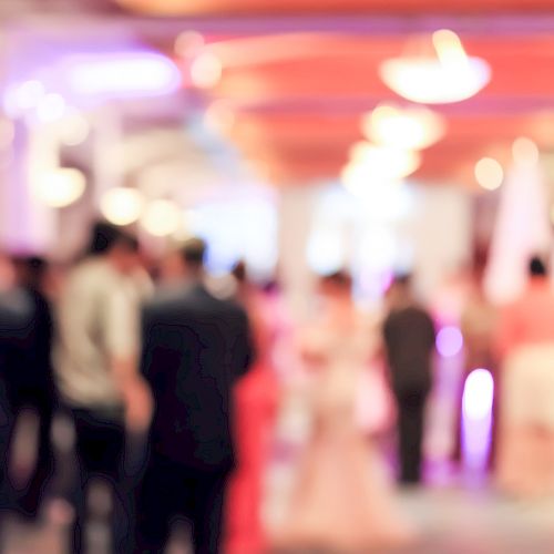 The image shows a blurred view of people attending a formal event or gathering in a well-lit, elegant venue.