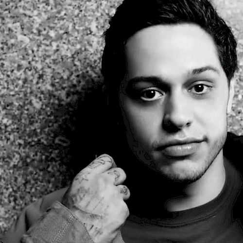 The image is a black-and-white photo of a person with short dark hair, looking at the camera, wearing a shirt and with tattoos on their hand, against a textured wall.