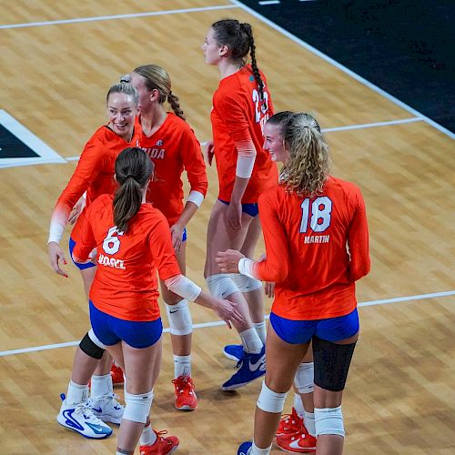 A group of female volleyball players wearing orange jerseys stand together on an indoor court, seemingly celebrating a point scored.