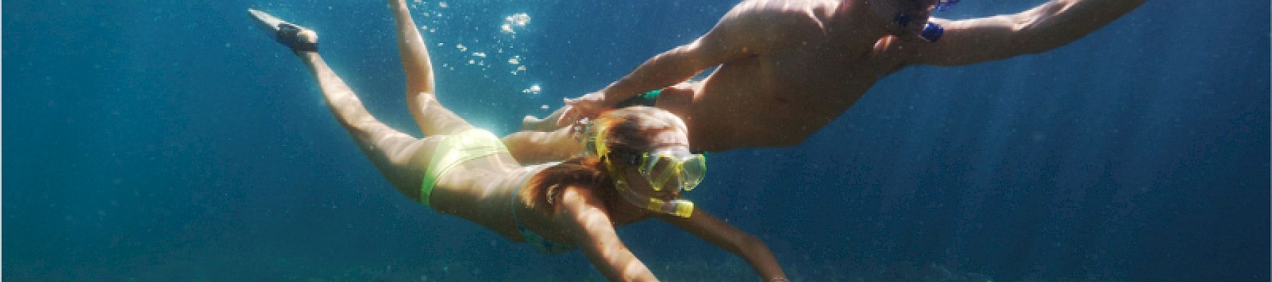 The image shows two people snorkeling underwater, swimming near rocks and coral, with clear blue water surrounding them.