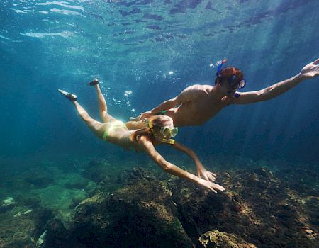 The image shows two people snorkeling underwater, swimming near rocks and coral, with clear blue water surrounding them.