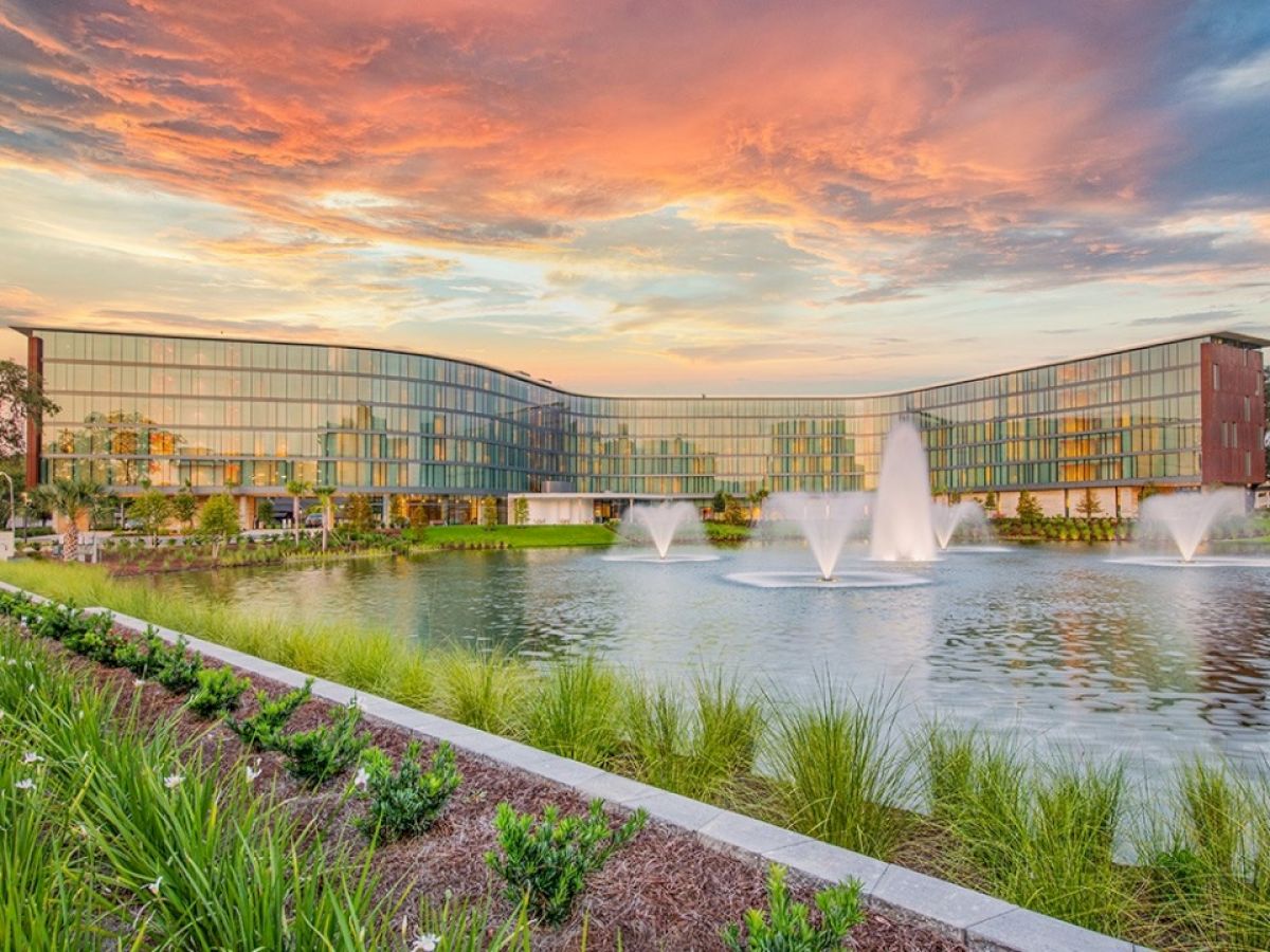 A modern building complex with a glass facade is mirrored in a pond featuring three large fountains, set against a vibrant and dramatic sunset sky.