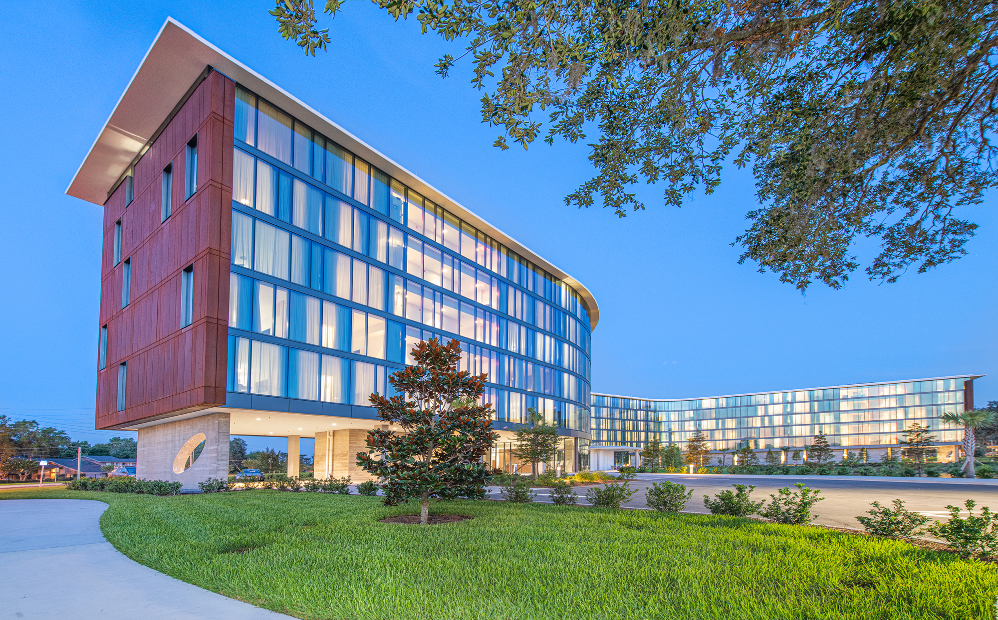 A modern, multi-story building with glass windows and a sleek design, surrounded by grass and trees under a clear blue sky.