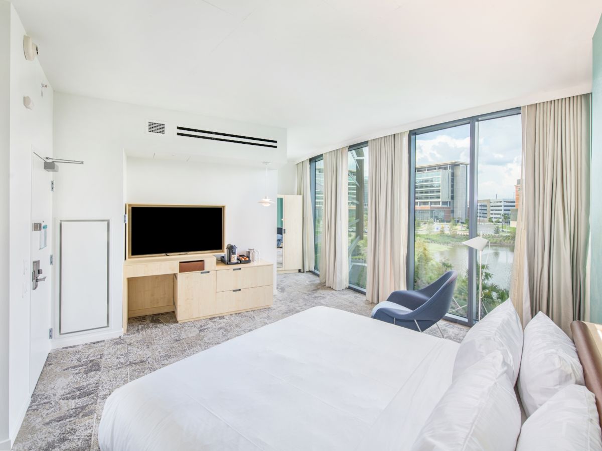 A modern hotel room with a large bed, flat-screen TV, armchair, and window with city view. Neutral tones and natural light fill the space.