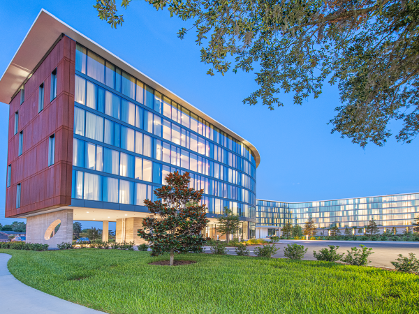 The image shows a modern, multi-story building with large glass windows, set against a clear blue sky, surrounded by landscaped greenery.