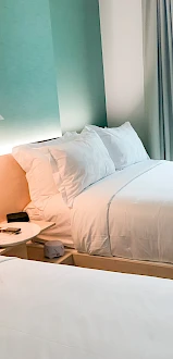 The image shows a hotel room with two neatly made beds, a bedside lamp, and light streaming in from a window with curtains.