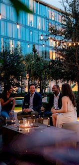People sitting outdoors on couches, surrounded by trees and string lights, with a modern building in the background.