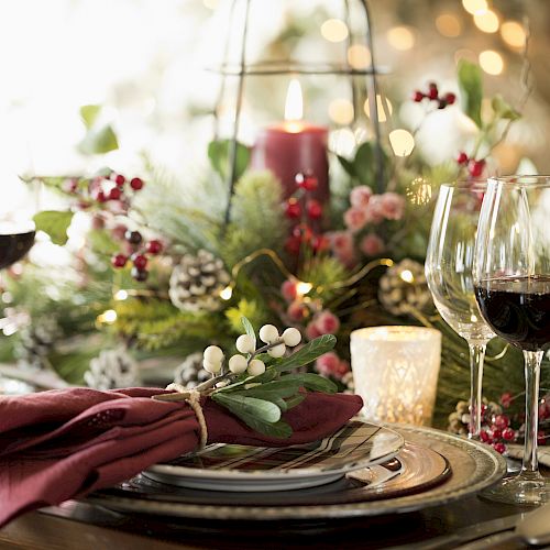 A festive table setting with wine glasses, pinecones, candles, berries, and greenery creates a warm holiday atmosphere.