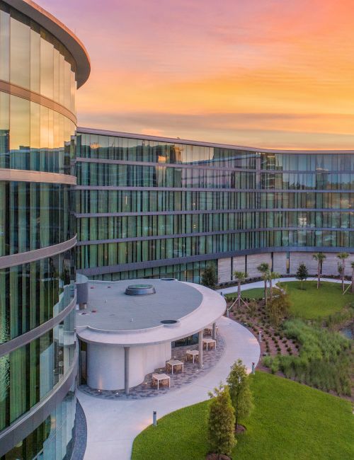 A modern glass building with a curved design overlooks a small pond, surrounded by greenery, under a vibrant orange and purple sunset sky.