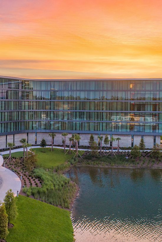 A modern glass building with a curved design overlooks a small pond, surrounded by greenery, under a vibrant orange and purple sunset sky.