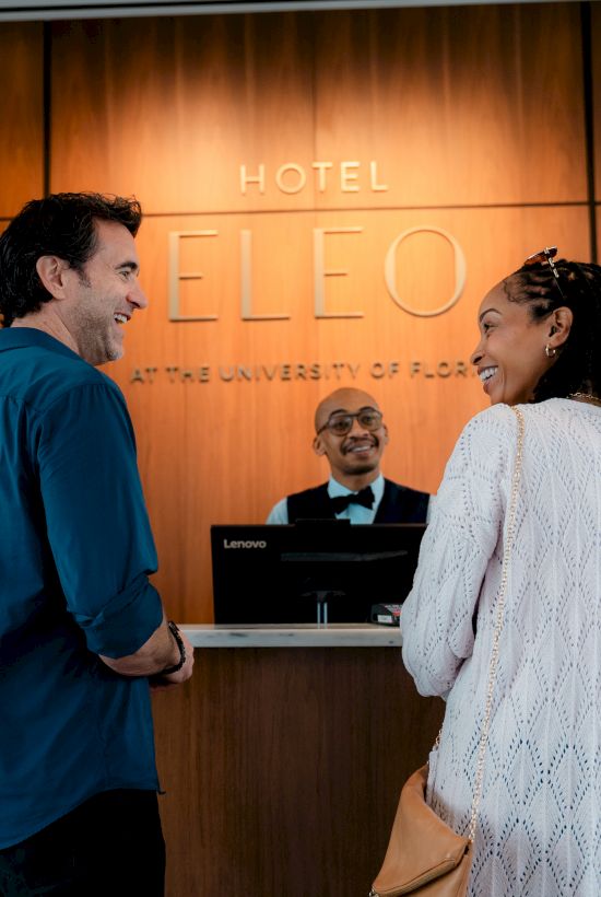 A couple is standing at a hotel reception desk, speaking to a receptionist. The ambiance is warm with flowers visible.