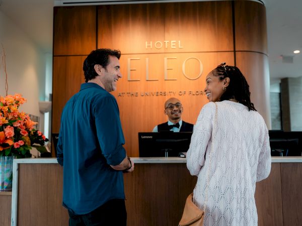 A couple is checking in at the reception of Hotel Eleo, University of Florida, greeted by a smiling receptionist.