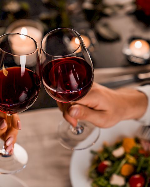 Two people clinking wine glasses over a candlelit dinner, with plates of salad visible, creating a romantic dining atmosphere.