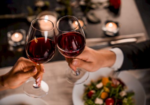 Two people clinking wine glasses over a candlelit dinner, with plates of salad visible, creating a romantic dining atmosphere.