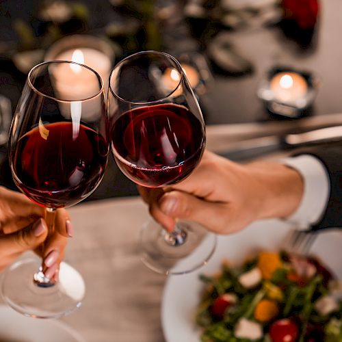 Two people clinking wine glasses over a candlelit dinner, with plates of salad visible, creating a romantic dining atmosphere.