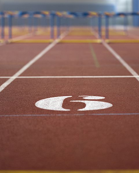 The image shows a track and field lane with the number 6, featuring hurdles in the distance.