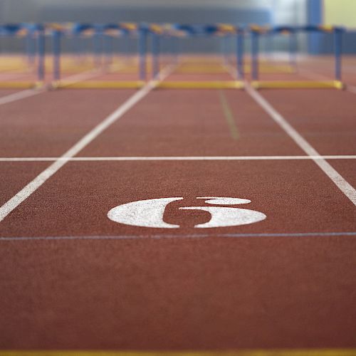 The image shows a track and field lane with the number 6, featuring hurdles in the distance.