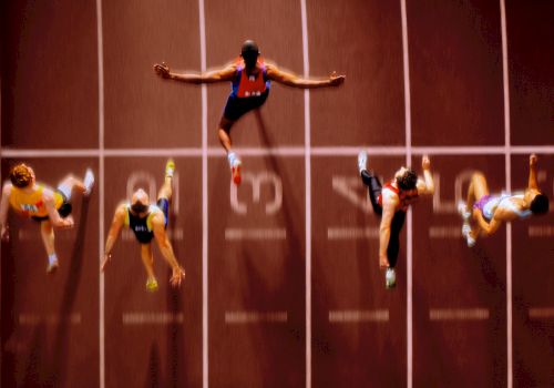 Aerial view of six sprinters crossing the finish line on a track, with arms outstretched and legs extended in mid-stride.