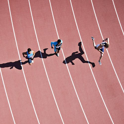 Three people are running on a track, casting long shadows on the surface, with one appearing to jump mid-stride.