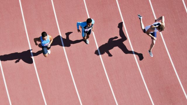 Three people are running on a track, casting long shadows on the surface, with one appearing to jump mid-stride.