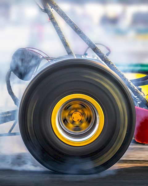 A close-up of a drag racing car's wheel, spinning and emitting smoke during a high-speed race, with a blurred background.