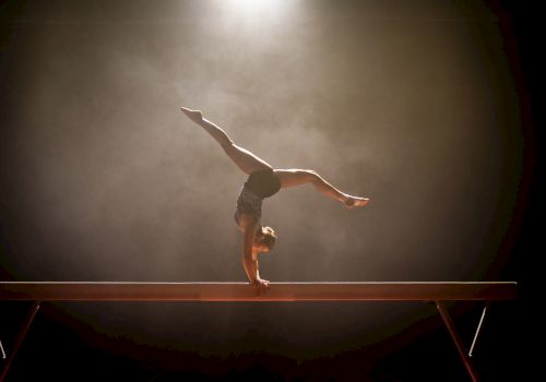 A gymnast performs a handstand on a balance beam, with a spotlight highlighting the scene against a smoky background.