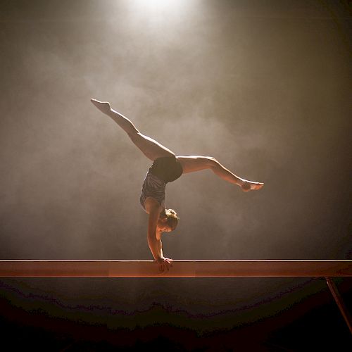 A gymnast performs a handstand on a balance beam, with a spotlight highlighting the scene against a smoky background.