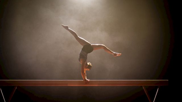 A gymnast performs a handstand on a balance beam, with a spotlight highlighting the scene against a smoky background.