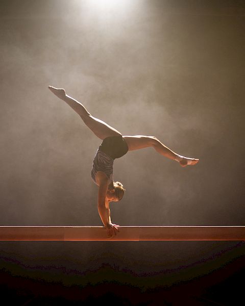 A gymnast performs a handstand on a balance beam, with a spotlight highlighting the scene against a smoky background.