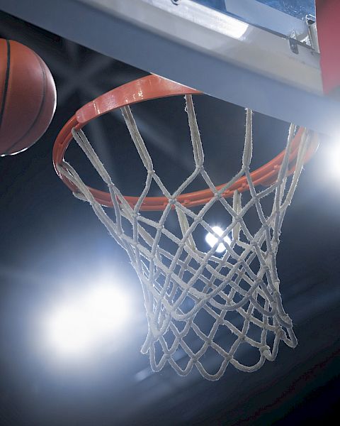 The image shows a basketball approaching a hoop, framed by bright stadium lights in the background.