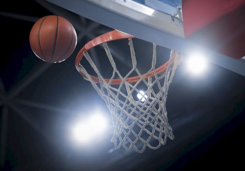 The image shows a basketball approaching a hoop, framed by bright stadium lights in the background.